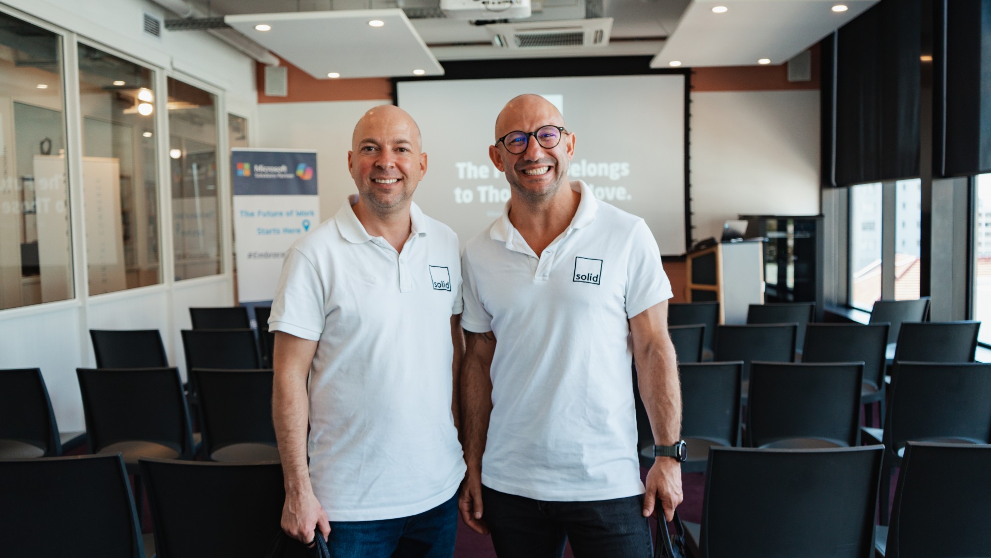 Co-founders Daniel Avinir and Michael Claxton, smiling in white Solid polo shirts, in a seminar room set up for a Solid masterclass