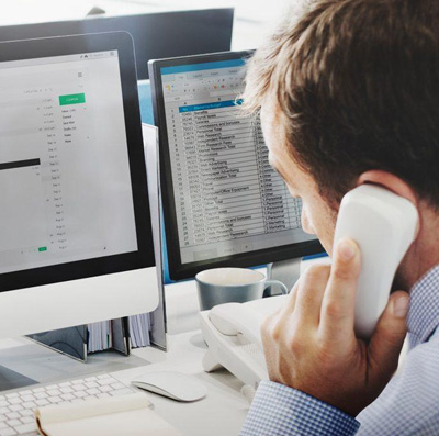 A man on the phone at a desk with two monitors showing email